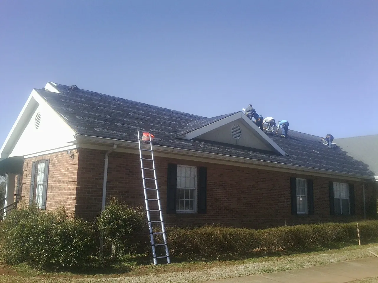 Crew tearing shingles off a brick ranch roof with a ladder set — Emergency Roof Repair work by our crew in La Crescent