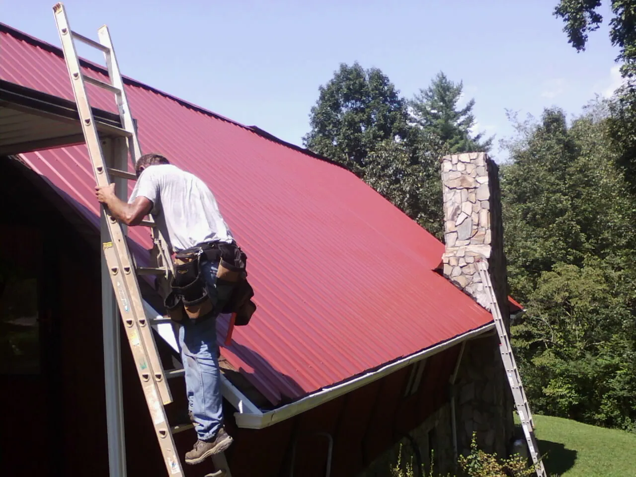 Roofer on a ladder beside a red metal roof and stone chimney — Soffit & Fascia Repair by Roofing Brothers in La Crescent